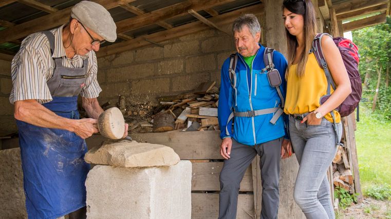 Un artisan sculpte un bloc de pierre dans un atelier. Deux visiteurs intéressés observent le processus.