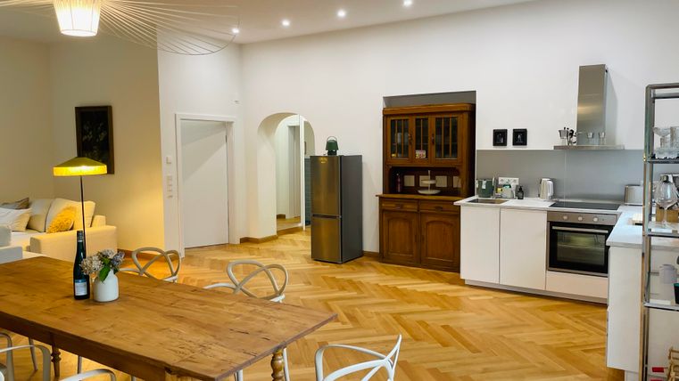 Une cuisine moderne avec un sol en bois et une grande table à manger. À l'arrière-plan se trouve un coin salon et un aménagement de cuisine.