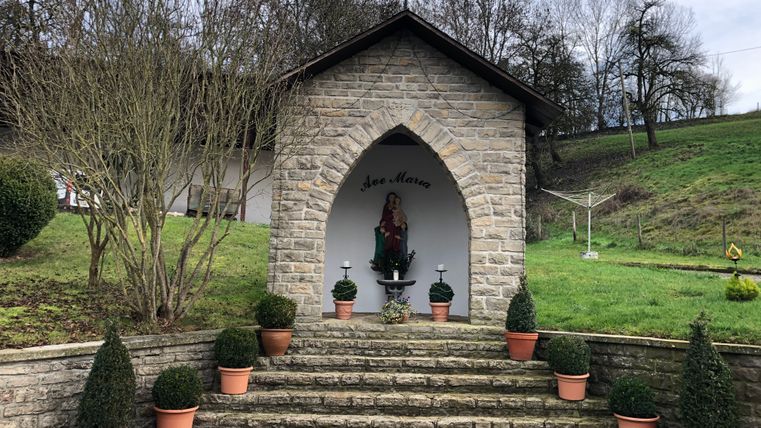Kleine steinerne Kapelle mit Marienstatue und Pflanzen in Töpfen.