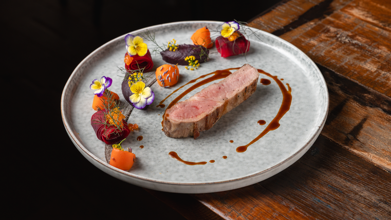 Une assiette artistiquement dressée avec un morceau de viande, garnie de légumes colorés et de fleurs comestibles. La présentation semble élégante et appétissante.