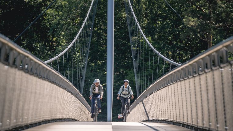Two cyclists on the Victor Neels Bridge, surrounded by trees.