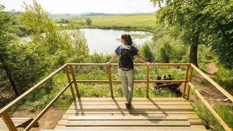 Wanderin steht auf einer hölzernen Plattform mit Sitzbank und blickt über das Geländer auf das Eichholzmaar, welches mitten in der grünen Landschaft liegt.