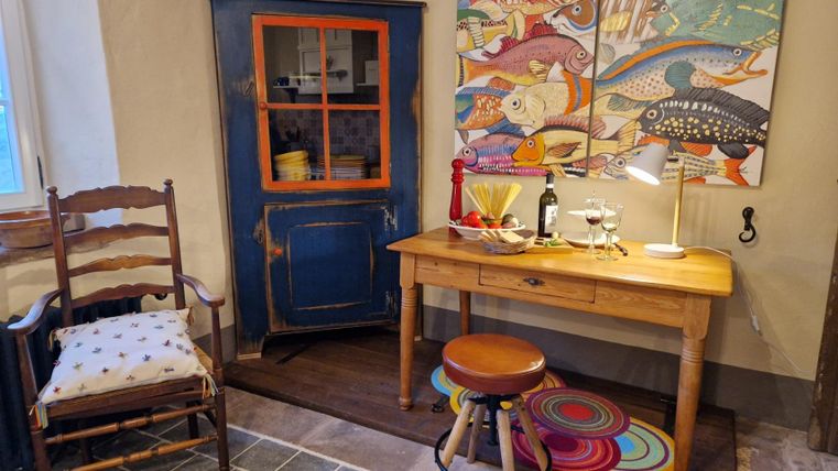 A cozy room with a wooden table, a lamp, and colorful decorations. In the background, there is a blue cabinet and a chair with a cushion.