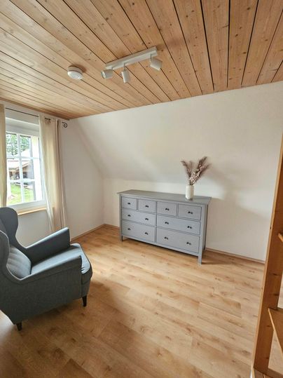 Een gezellige kamer met houten plafond, een blauwe fauteuil en een grijze commode. Het licht valt door een raam en creëert een uitnodigende sfeer.