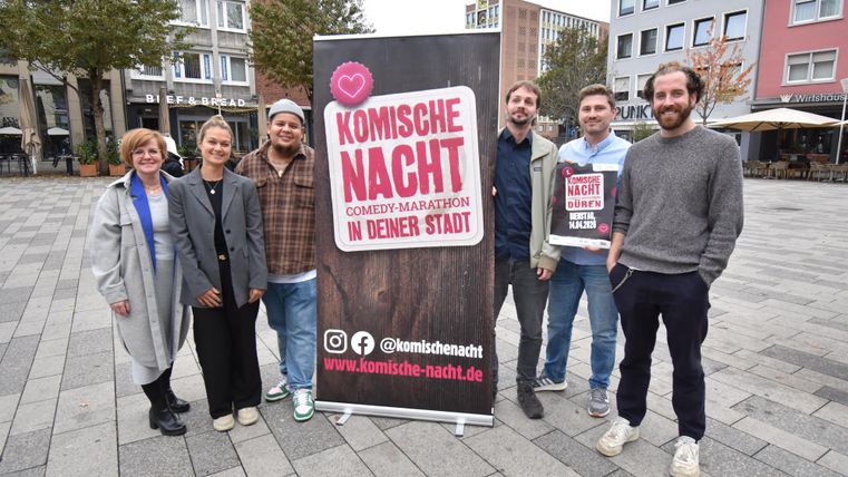 Eine Gruppe von sechs Personen steht vor einem Banner, das für eine Comedy-Veranstaltung mit dem Titel "Komische Nacht" wirbt. Die Szenerie ist urban, mit modernen Gebäuden im Hintergrund.