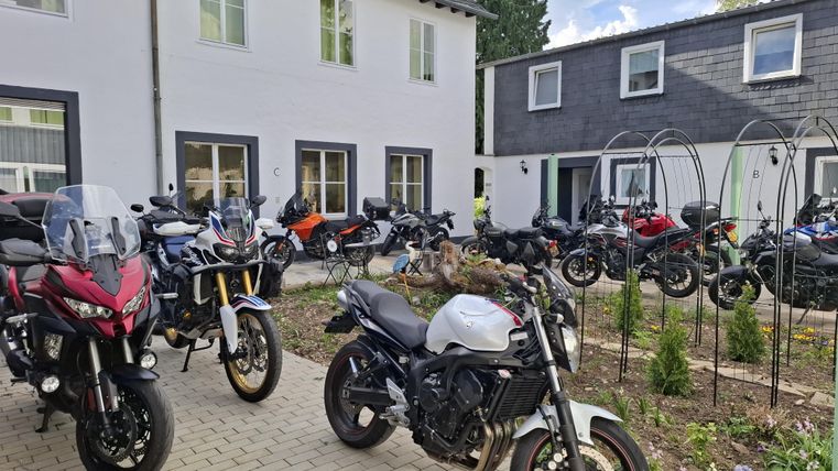 Eine Gruppe von Motorrädern steht auf einem gepflasterten Hof vor einem zweigeschossigen Gebäude. Im Hintergrund sind weitere Motorräder und ein gepflegter Garten zu sehen.