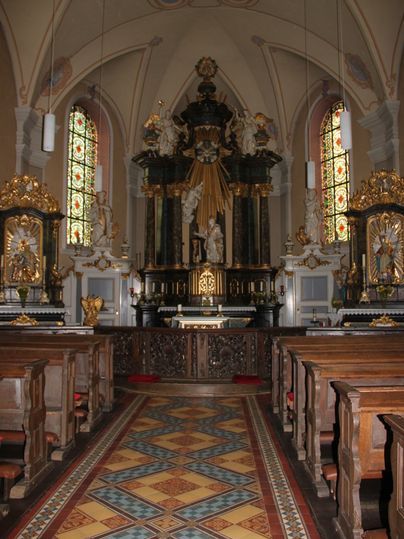 Interieur van een kerk met altaar, kerkbanken en kleurrijke ramen.