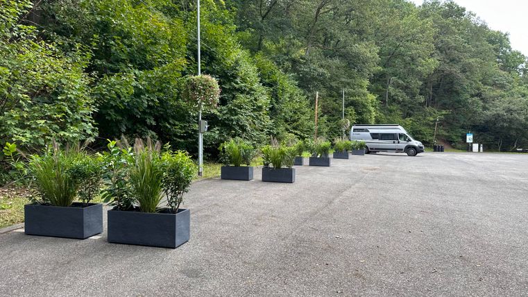 Ein Parkplatz mit großen Pflanzkästen und dichter, grüner Vegetation im Hintergrund. Ein Transporter steht am Rand des Parkplatzes.