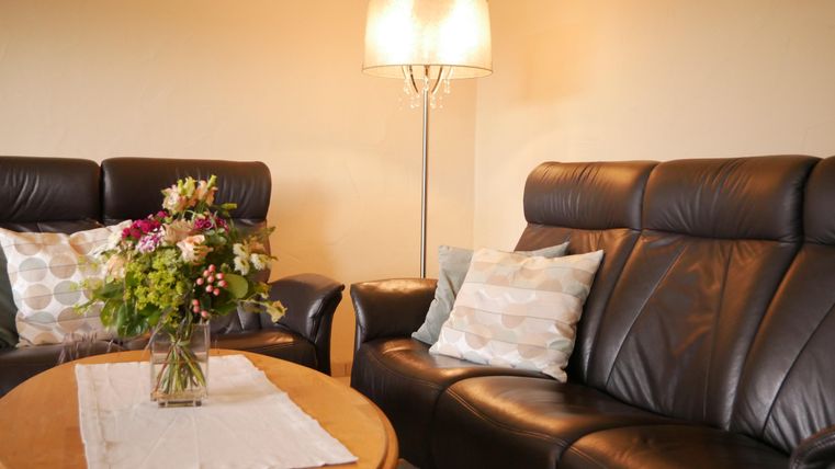 Een gezellige woonkamer met twee zwarte leren banken en een ronde houten tafel. Op de tafel staat een vaas met kleurrijke bloemen en een tafellamp zorgt voor warm licht.