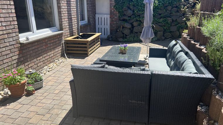 Een gezellige terras met een rietenachtige bank en een kleine tafel. Op de achtergrond zijn planten en een parasol te zien.