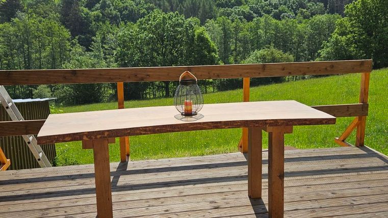 A wooden table stands on a terrace overlooking the green valley and the trees. The sky is clear and sunny, which enhances the peaceful atmosphere.