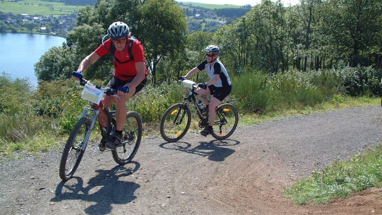 Twee mountainbikers fietsen een steile weg omhoog. Op de achtergrond is een mooi meer en een groene landschap te zien.