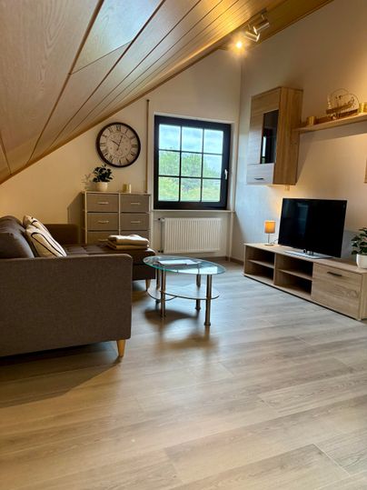 A modern living room with slanted walls and a large window. There is a sofa, a coffee table, and a TV in a bright, cozy room.