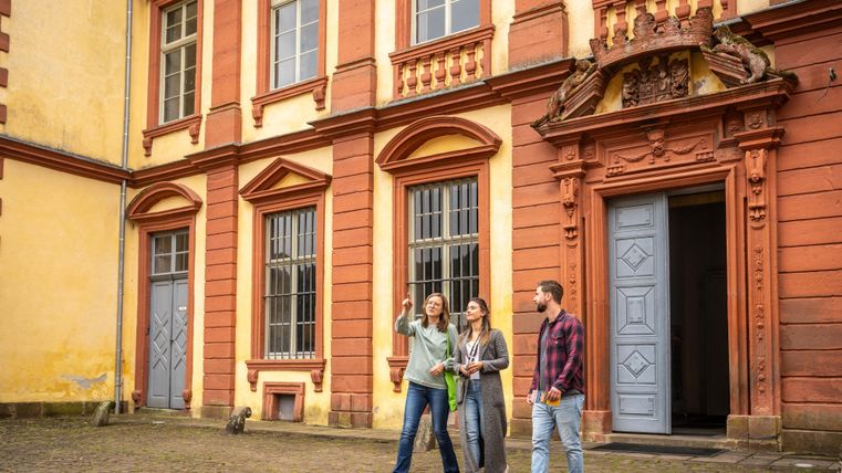 Drie personen staan voor een historisch gebouw. Een vrouw wijst naar iets, terwijl de anderen geïnteresseerd luisteren.