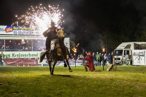 A rider on a horse is performing a spectacular display with fireworks in the background. Spectators are watching the event at night.