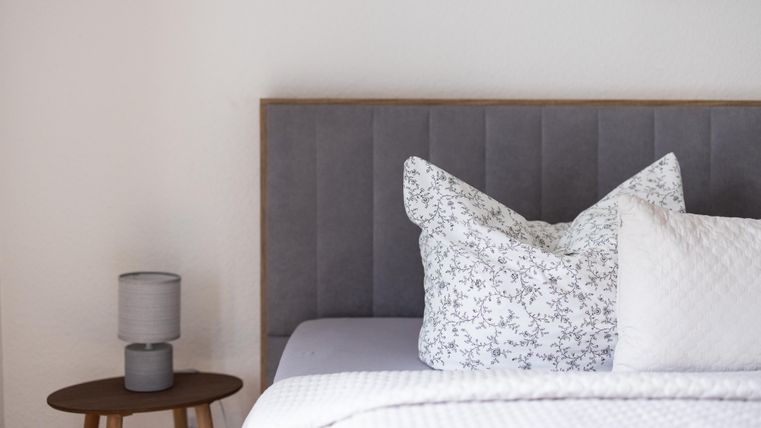 Een moderne slaapkamer met een grijze hoofdbord en elegante kussens. Naast het bed staat een kleine tafel met een lamp.
