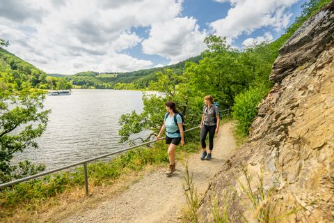 Zwei Wandererinnen auf einem schmalen Pfad am Rursee, der sich entlang eines felsigen Hangs schlängelt. Umgeben von üppigem Grün und bewaldeten Hügeln genießen sie die Aussicht auf das ruhige Wasser unter einem teils bewölkten Himmel