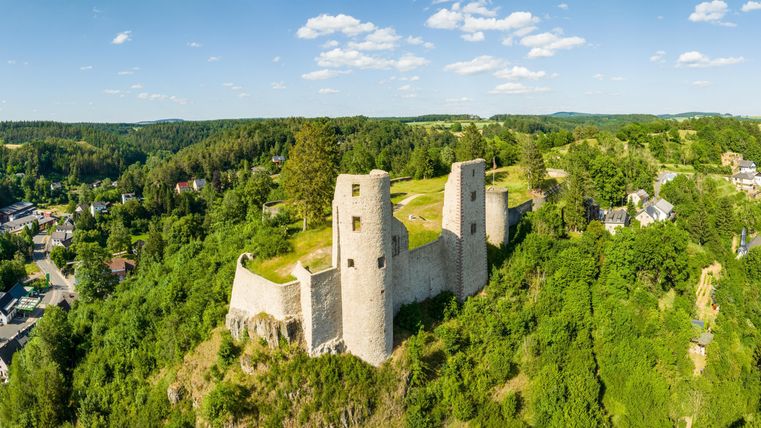 Une vieille ruine de château sur une colline, entourée d'arbres verts. Le ciel est bleu avec quelques nuages.