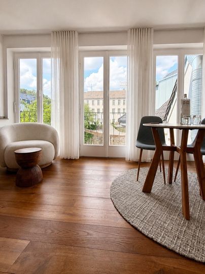 Een moderne woonkamer met grote ramen en een mooi uitzicht naar buiten. De ruimte is ingericht met een rond tapijt, een kleine tafel en elegante meubels.