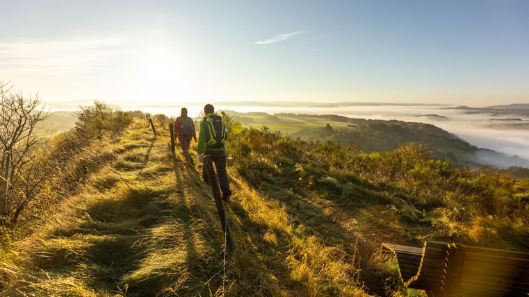 Two hikers are walking along a path through a hilly landscape. In the background, a clear sky and a bank of fog can be seen.