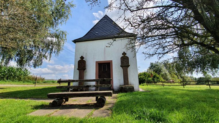 Een kleine witte kapel met een leistenen dak, omringd door bomen en een bankje op de voorgrond.