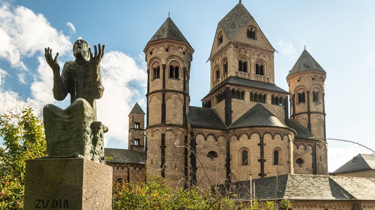 Een standbeeld van een biddende persoon staat voor een indrukwekkende kerk met torens. De lucht is blauw en er zijn enkele wolken.