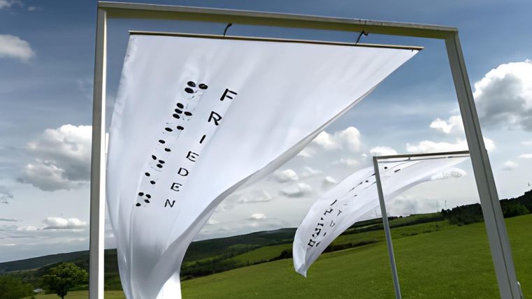 Kunstinstallation bestehend aus zwei Stahlkonstruktionen, an denen weiße Fahnen mit dem Schrifzug "Frieden" angebracht sind auf einer großen Wiese.
