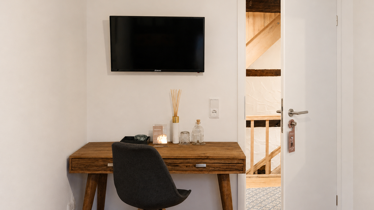 Une chambre moderne avec une table en bois et une télévision sur le mur. La porte mène à une autre pièce avec des structures en bois visibles.
