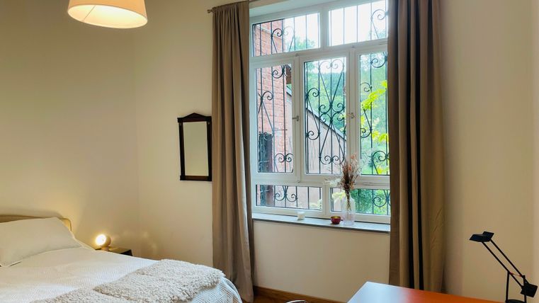 Une chambre lumineuse avec une grande fenêtre et des rideaux modernes. Sur le côté gauche se trouve un lit avec une couverture confortable et un bureau avec une lampe.