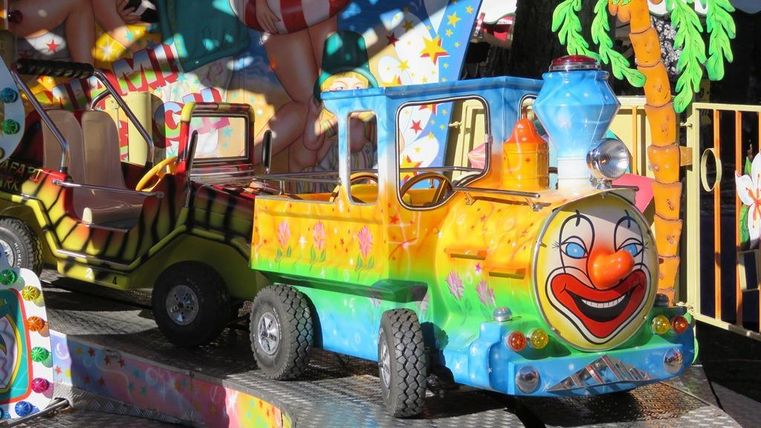 Colorful rides at a fair with a clown face. Some palm trees and vibrant colors give the image a cheerful atmosphere.