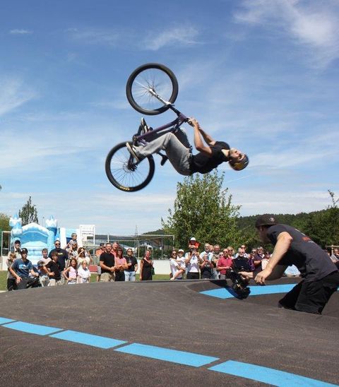 Ein BMX-Fahrer macht einen spektakulären Rückwärtssalto über eine Rampe. Im Hintergrund versammelt sich eine enthusiastische Menge, die die Aufführung verfolgt.