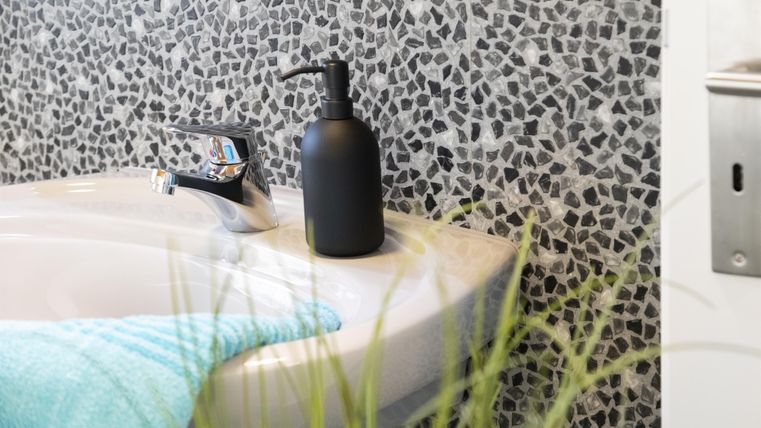 Een moderne wastafel met een zwarte zeepdispenser en een handdoek in turquoise. Op de achtergrond is een gestructureerde muur met kleine stenen te zien.