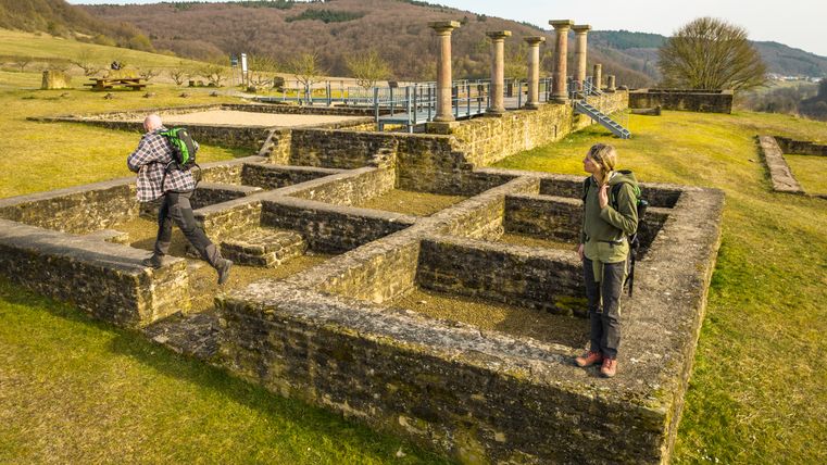 Twee mensen verkennen de ruïnes van de Romeinse Villa Holsthum met zuilen en stenen muren op de achtergrond.