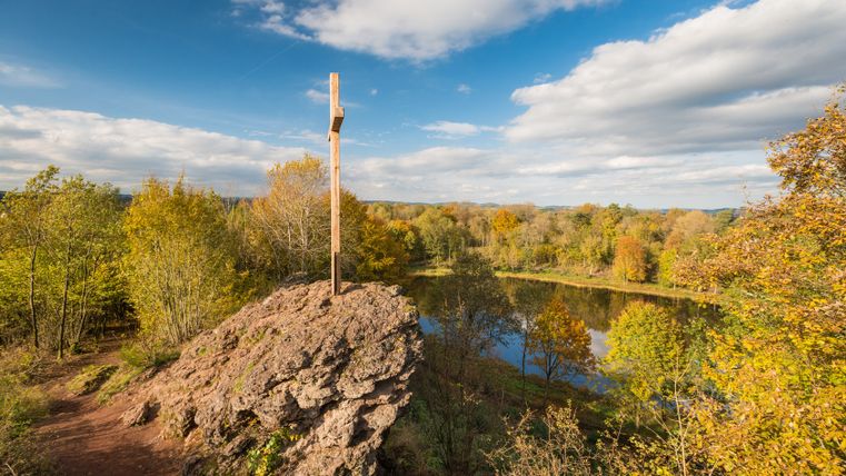 Ein Holzkreuz auf einem Felsen überblickt einen Kratersee, umgeben von herbstlichen Bäumen unter einem blauen Himmel mit Wolken.