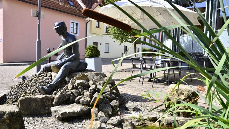 Bronze statue of a seated man on stones, surrounded by reeds and a small pond, with houses and a parasol in the background.