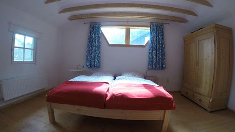 Ein helles Schlafzimmer mit einem großen Bett und roten Bettdecken. Es gibt ein Fenster mit blauen Vorhängen und einen Holzschrank im Raum.