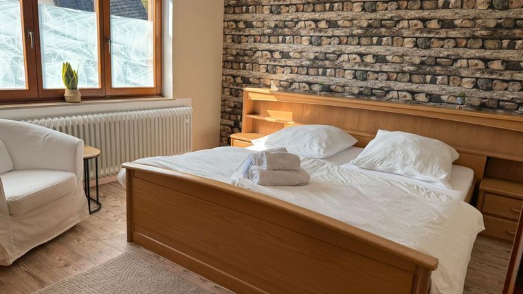 A cozy bedroom with a double bed and fresh white bed linens. The room has large windows and a beautiful stone wall.