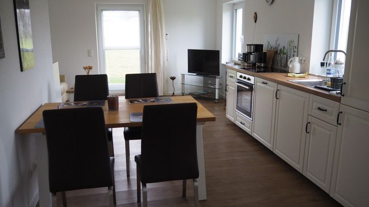 Eine helle Küche mit einem Esstisch und Stühlen. Im Hintergrund sind ein Fernseher und ein Fenster mit Blick nach draußen sichtbar.