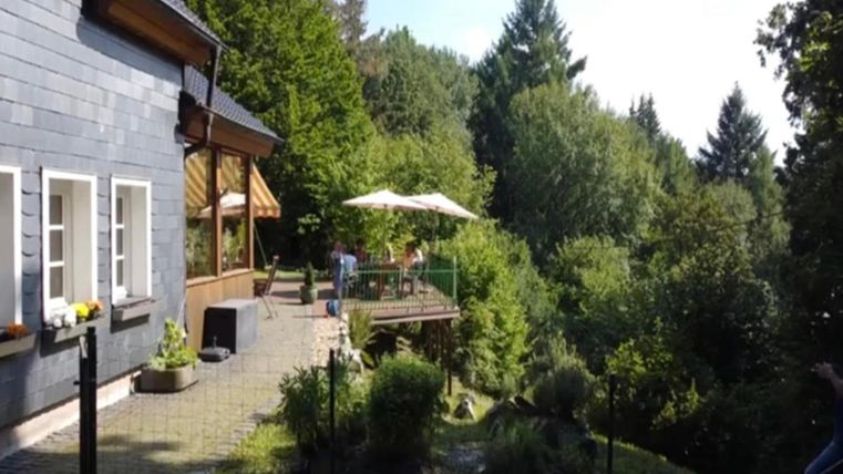 A cozy house surrounded by trees and greenery. In the background, there is a terrace with umbrellas and chairs visible.