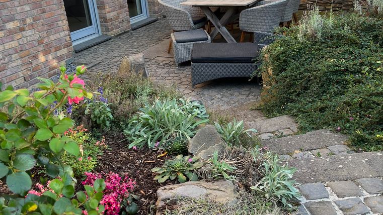 A cozy outdoor area with a table and chairs. Colorful flowers surround the seating area, and a stone wall forms the backdrop.