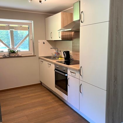 Une cuisine moderne avec des armoires blanches et des détails en bois. Elle est bien équipée avec une cuisinière, une bouilloire et une fenêtre qui laisse entrer la lumière du jour.