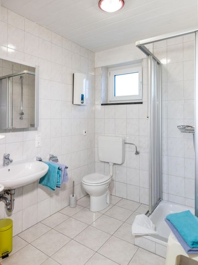 Een heldere, moderne badkamer met een douche, wastafel en toilet. De muren zijn in lichte tegels gehouden, en er zijn enkele handdoeken in felle kleuren.