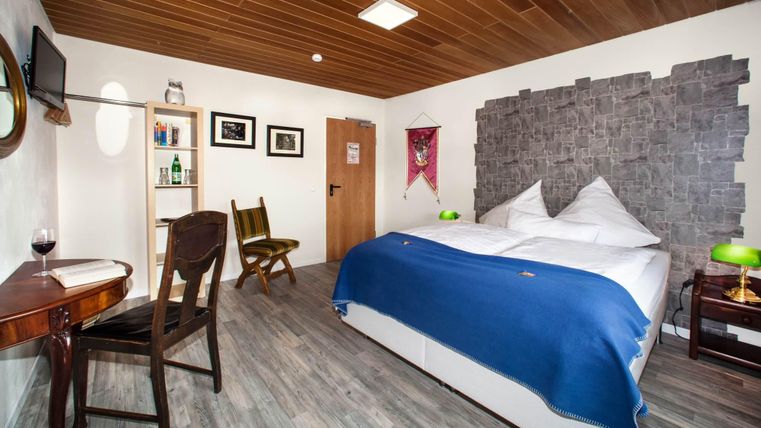 A modern hotel room with a large bed and blue bed linen. There is a table, a television, and a pleasant wooden ceiling.