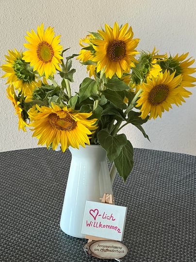 Een boeket zonnebloemen in een witte vaas staat op een tafel. Daaronder bevindt zich een welkomstbord.