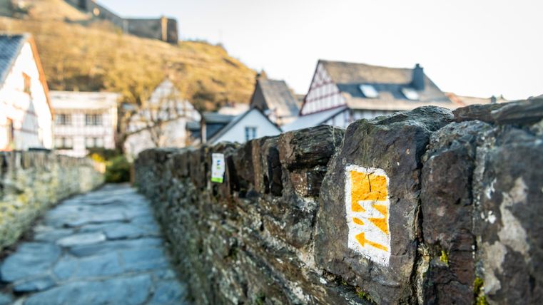 Wandelpadmarkering op een stenen muur met vakwerkhuizen op de achtergrond.