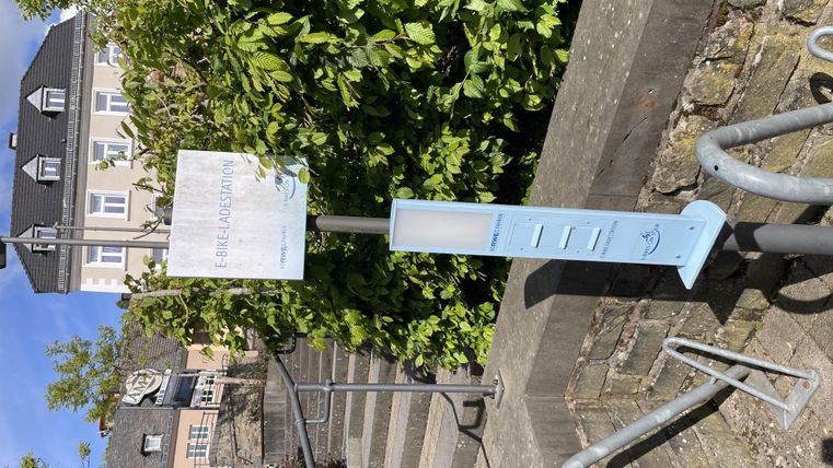 An information pillar stands at a staircase, surrounded by green plants. In the background, some houses can be seen.