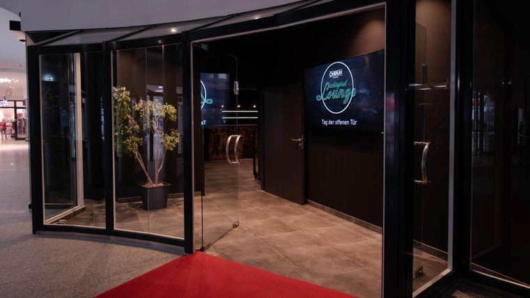 Ein stilvoller Eingangsbereich mit gläsernen Türen und einem roten Teppich. Im Inneren sind moderne Bildschirme und eine gemütliche Atmosphäre zu sehen.