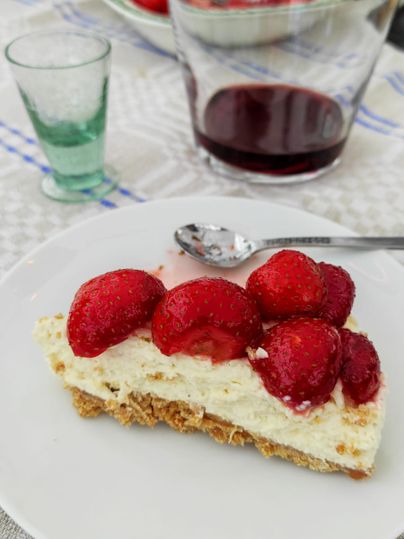 Ein Stück Erdbeerkuchen auf einem weißen Teller, daneben ein Glas und eine Karaffe mit rotem Getränk auf einem Tisch.