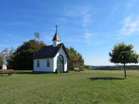Een kleine witte kapel met een groene poort en een kruis op het dak staat op een weiland onder een blauwe hemel.