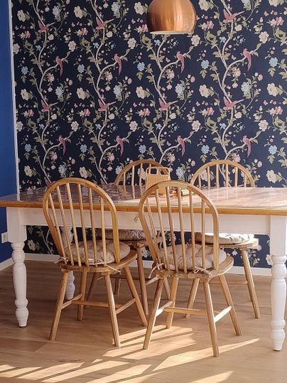A cozy dining area with a wooden table and four chairs. The walls are adorned with a blooming, dark wallpaper pattern.